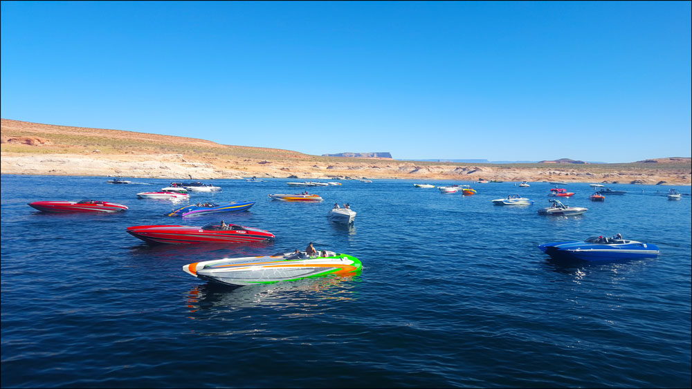 Colorful speedboats drifting on calm blue water near a desert shoreline under a clear sky, conveying a peaceful, recreational atmosphere.